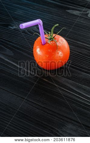 One red tomato with straw on black wood background. Fresh juice healthy food concept. Tomato as a vessel with fresh juice. Background with a Blank Space for Text