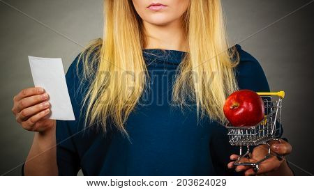 Worried woman holding shopping basket with fruits looking at bill receipt being scared of huge prices
