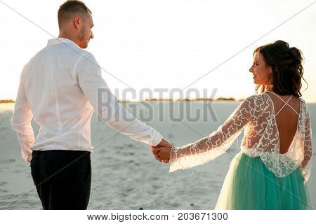 Bride and groom go on sand in desert hold hands and look at each other. They go on background of white sand. Back view. Close up.