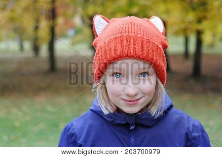 Cheerful girl smiling, the child is dressed in a funny knitted warm hat with ears, looks like a fox. Autumn, outdoors portrait.
