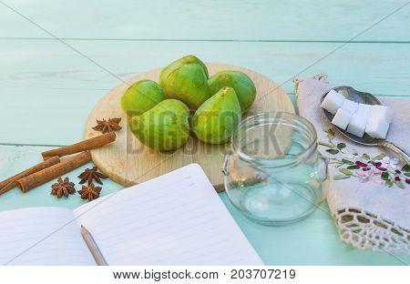 Still life with figs, jam cooking process. Fresh green figs on wooden backround, surrounded by spices, embroidered nupkin, vintage spoon, a glass container for jelly. Notebook with copy space.