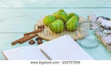 Still life with figs, jam cooking process. Fresh green figs on wooden backround, surrounded by spices, embroidered nupkin, vintage spoon, a glass container for jelly. Notebook with copy space.