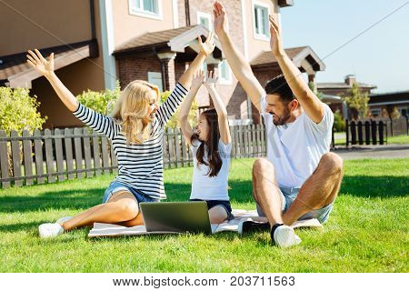Upsurge of joy. Happy lovely family sitting on the lawn in their backyard and raising hands up, cheering happily