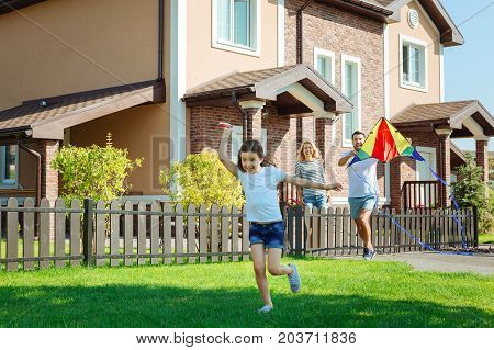 Amusing pastime. Charming little girl running across backyard of her house and flying a kite with her parents