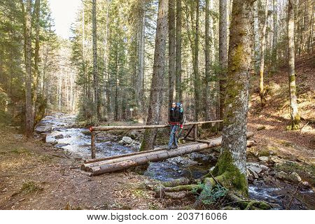 Woman hiker crossing a footbridge in forest across a pure mountain river with a blaze drawn for direction sun shining. Active lifestyle pristine nature concept.