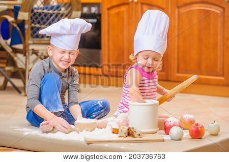 Funtime: two siblings - boy and girl - in chef's hats sitting on the kitchen floor soiled with flour playing with food making mess and having fun