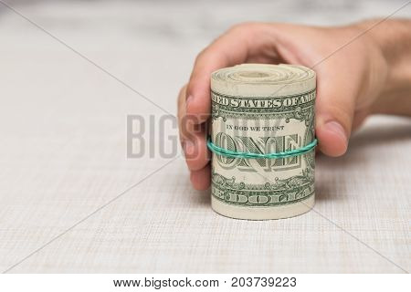 A man holds a bunch of money twisted in a bunch and tied with a green rubber band on a light textured background