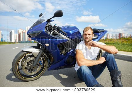 Rear view of unrecognizable Caucasian man motorbiker wearing jeans leather jacket and boots standing next to his blue parked custom-built motorcycle and carrying helmet. Male with chopper outdoors
