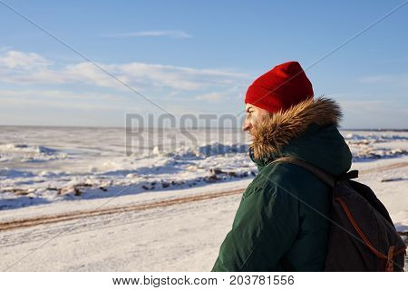 Handsome bearded man wearing red hat and coat with fur hood carrying backpack hitchhiking on road in rural area while traveling alone in winter. People lifestyle travel and trekking concept