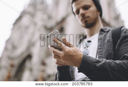 Cut view of a young tourist standing near the tall church building. He is searching some information on the Internet by using his phone. Close up