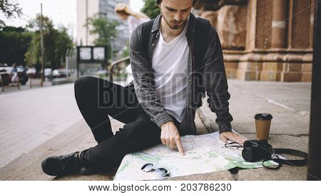 Close view of a young hipster that likes to travel. He decided to have some rest and sit down on the cathedral's steps on the street. His coffee is getting colder while the guy is stading the map of the city. Close up