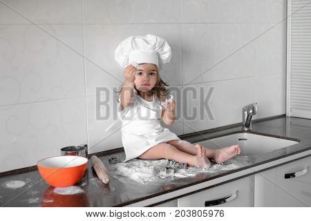 Little smiling baby girl baker in white cook hat and apron kneads a dough on tle kitchen.