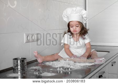 Little smiling baby girl baker in white cook hat and apron kneads a dough on tle kitchen.