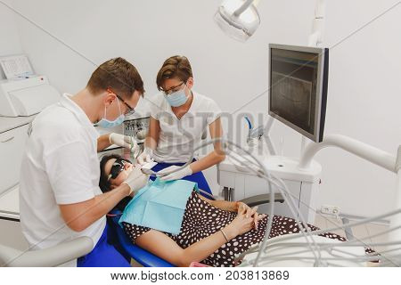 A Male Dentist With A Female Assistant Helps To Treat The Teeth Of A Woman Patient In A Clinic In Th