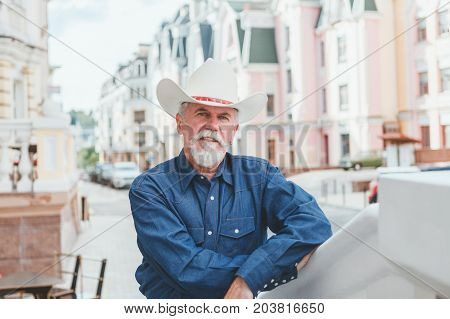 A mature cowboy in a hat jeans and a denim shirt looks at the camera. On open air