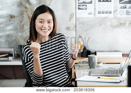 Young businesswoman with arms up and smiling face while working with laptop computer at her office table success in business concept