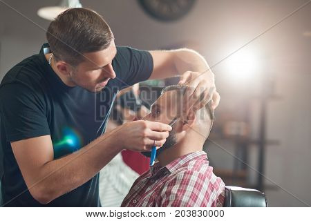 Professional barber working shaving his bearded client at the barbershop.