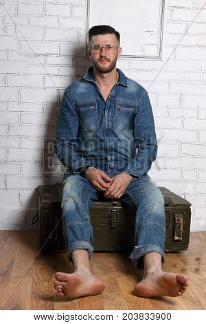 Barefoot man in glasses and denim overalls sits on wooden box in white room