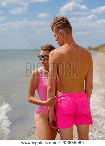 The woman is behind the man. Natural and sea view on the background. Joyful and romantic couple on sunset, at the golden beach, blue sky on the background. Sunny day outside. Love is in the air.