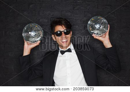 Young brunette smiling man wearing black suit white shirt sunglasses with black frames holding two disco balls.
