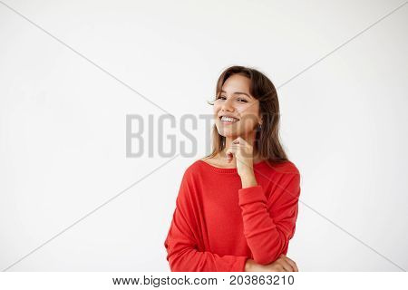 Positive human facial expressions and emotions. Isolated shot of attractive European student girl in red jamper looking and smiling broadly at camera during nice conversation with someone