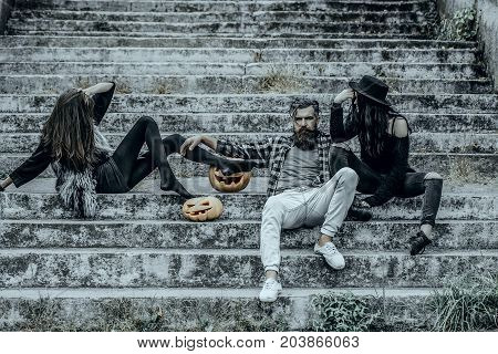Halloween friends on stony stairs. Women with pumpkin heads. Bearded man sitting with girls. Halloween people and jack o lanterns. Holiday celebration concept.