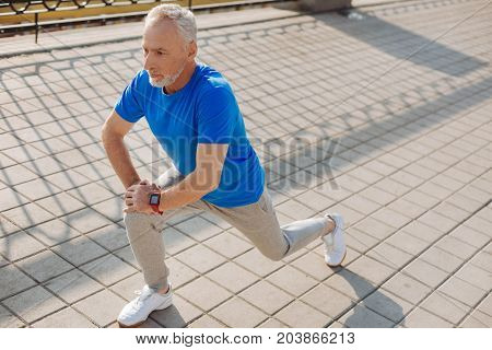Energetic warm-up. Serious fit senior man warming up before his morning run and doing lunges as a part of his stretching routine