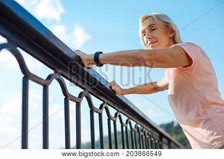Necessary rest. Pleasant elderly woman holding her hands on the balustrade of the bridge and regaining her breath after a morning run