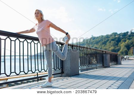 Supple muscles. Charming senior woman holding on to the bridge balustrade and performing leg stretching before her morning run