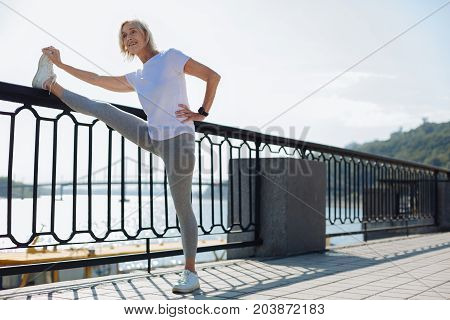 Excellent suppleness. Upbeat athletic senior woman holding her leg on the top of a bridge balustrade while performing stretching exercises and demonstrating good suppleness