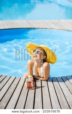 Young woman in swimsuit with big yellow sunhat relaxing with a bottle of fresh drink sitting on the poolside outdoors