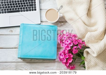 Female workplace with a laptop cup of coffee blue notebook cozy sweater and pink flowers on light wooden background.Top view composition with copy space.