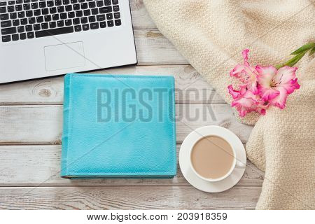 Female workplace with a laptop cup of coffee blue notebook cozy sweater and pink flowers on light wooden background.Top view composition with copy space.