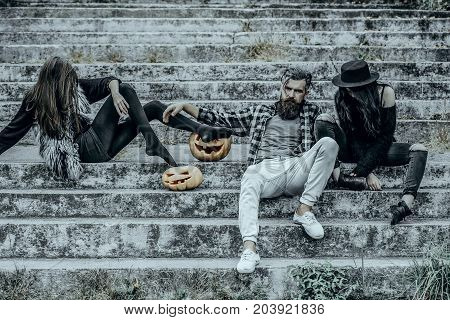 Halloween holiday celebration concept. Bearded man sitting with girls. People or friends on stony stairs. Halloween jack o lantern symbols. Women with pumpkin heads.