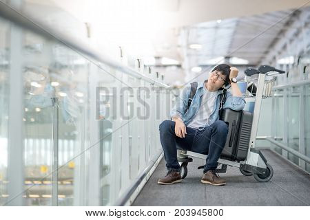 Young Asian man sitting on airport trolley with his suitcase luggage in the international airport terminal waiting for travel abroad