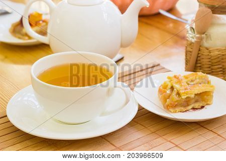 White cup with a herbal tea and a piece of cake on a saucer on the bamboo napkin on a blurred background of a teapot and other tea appliances
