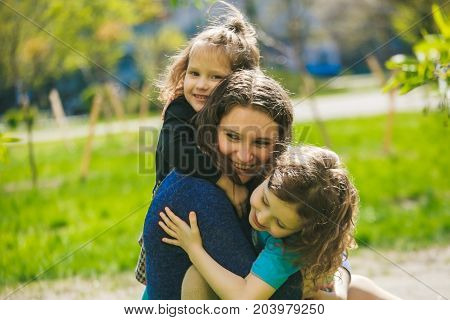 Little girl hugs mother sitting on the grass. Child playing threw his mother on grass. Family in spring walks on street.