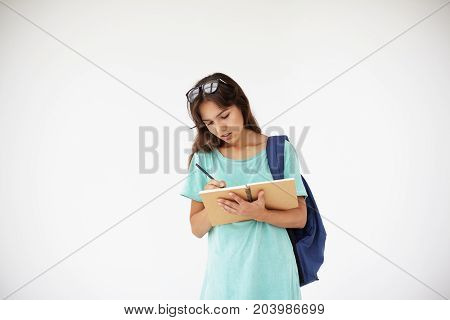 People youth education learning and knowlegde concept. Picture of beautiful college student girl wth glasses on head carrying backpack and writing down in copybook posing isolated in studio