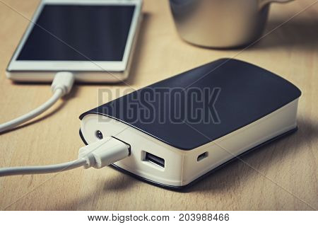 Closeup of a powerbank charging a smartphone on a wooden table next to a cup of coffee