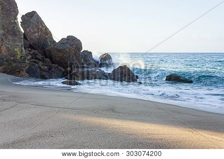 Breaking Waves Splashing On Cliff Face With Rocks And Foam Backwash On Sandy Shore