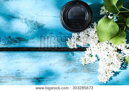 Black Cardboard Cup Of Coffee And A Branch Of Lilac On A Blue Wooden Bench Against Green Grass And S