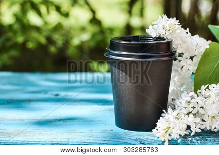 Black Cardboard Cup Of Coffee And A Branch Of Lilac On A Blue Wooden Bench Against Green Grass And S