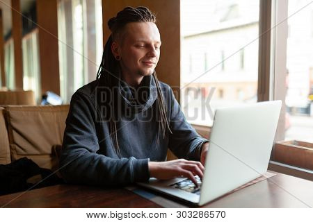 Young Businessman With Dreadlock Having Doing His Work In Cafe Near The Big Window With Laptop. Yout