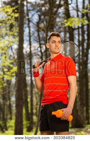 The Man In A Red T-shirt Pose With A Tennis Racket And Orange Thermocouple, On The Background Of Gre