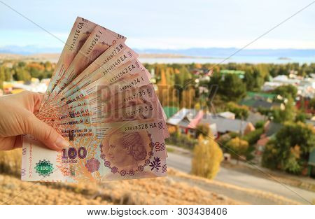 Woman Hand Holding Bunch Of Argentinian 100 Pesos Banknotes (front) With Blurry El Calafate Town In 