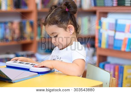 Little Child Indoors In Front Of Books. Cute Young Toddler Sitting On A Chair Near Table And Play Wi