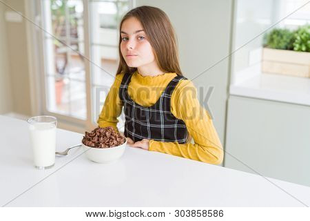 Beautiful young girl kid eating chocolate cereals and glass of milk for breakfast with serious expression on face. Simple and natural looking at the camera.