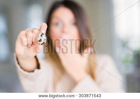 Young beautiful woman holding fresh quail egg at home cover mouth with hand shocked with shame for mistake, expression of fear, scared in silence, secret concept