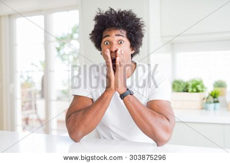 Young african american man wearing casual white t-shirt sitting at home afraid and shocked, surprise and amazed expression with hands on face