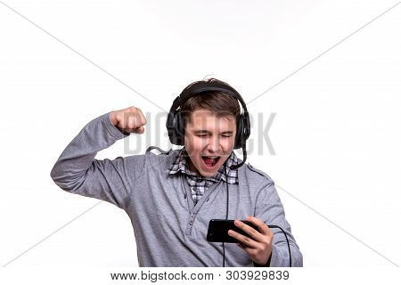 Schoolboy Guy With Headphones Playing On Mobile Phone. Teenager Happy Screams . Young Man Won An Onl